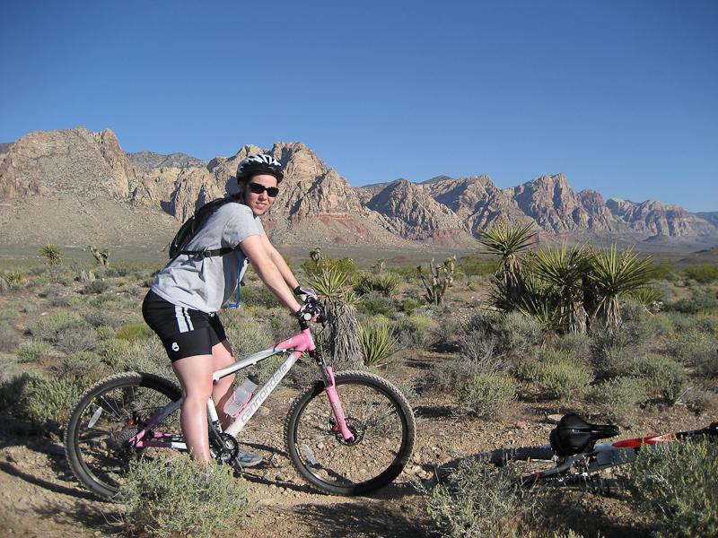 A person riding a mountain bike in a desert landscape with rocky mountains in the background. The cyclist is wearing a helmet and sunglasses, dressed in a gray shirt and black shorts, with a backpack on. There are sparse shrubs and desert vegetation surrounding the area. Blue Diamond mountain bike trail.