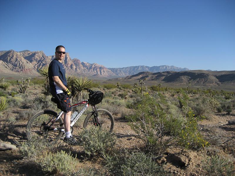 A person standing next to a mountain bike in a desert landscape, surrounded by sparse vegetation and rocky mountains in the background under a clear blue sky. The individual is wearing casual biking attire and gloves, looking towards the camera. Blue Diamond mountain bike trail.