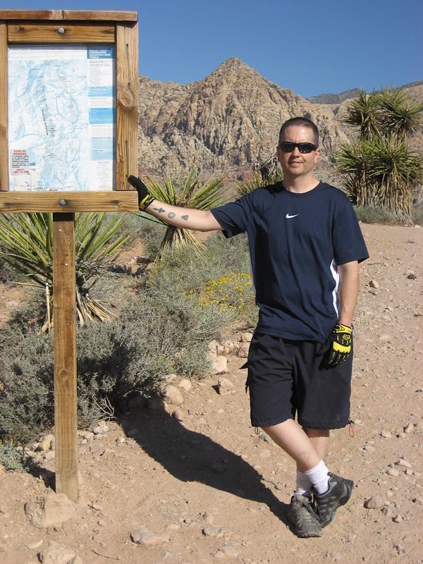 A man in a black Nike athletic shirt and shorts stands next to a wooden information sign in a mountainous outdoor setting. He is wearing yellow and black gloves and sunglasses, and there are desert plants in the background. The sun is shining, indicating a bright, clear day. Blue Diamond mountain bike trail.