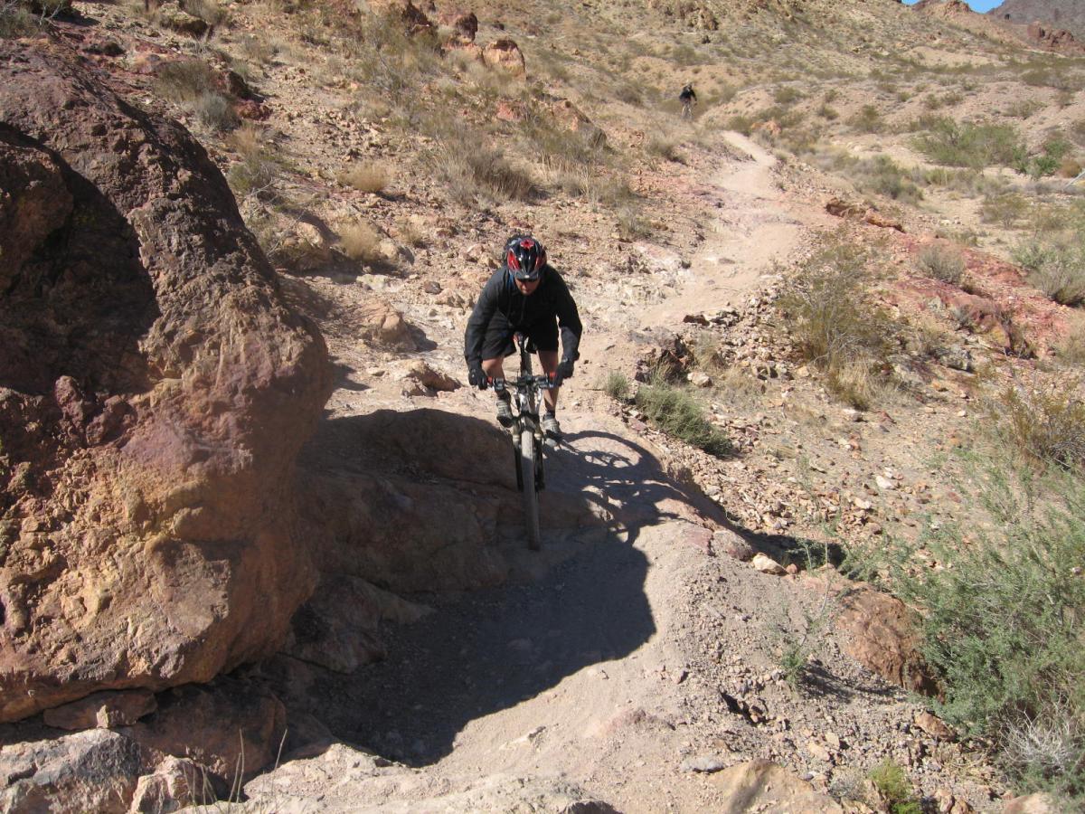Mountain biker navigating a rocky trail in a desert landscape, with shrubs and small hills in the background. The cyclist is wearing a helmet and biking gear while maneuvering over uneven terrain. Bootleg Canyon mountain bike trail.