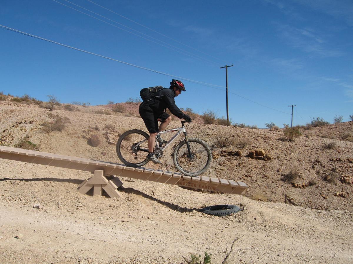A mountain biker riding along a narrow wooden bridge over a dry, rocky landscape. The cyclist is wearing a helmet and is in motion, navigating the bridge with power lines visible in the background. Bootleg Canyon mountain bike trail.