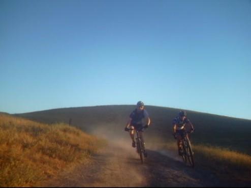 Two mountain bikers riding along a dusty dirt trail in a scenic outdoor setting, with rolling green hills and a clear blue sky in the background. Briones Regional Park mountain bike trail.