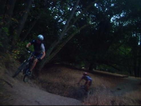 Two mountain bikers navigating a dirt trail surrounded by trees and underbrush. One biker is in the foreground, jumping off a small rise, while the other is riding in the background. The scene is set in a wooded area, showcasing a rugged terrain. Briones Regional Park mountain bike trail.