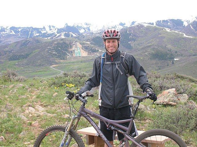 A smiling mountain biker wearing a helmet stands beside his bike on a scenic mountain trail. The background features lush green hills and snow-capped peaks under a cloudy sky, showcasing a beautiful outdoor landscape. Glenwild mountain bike trail.