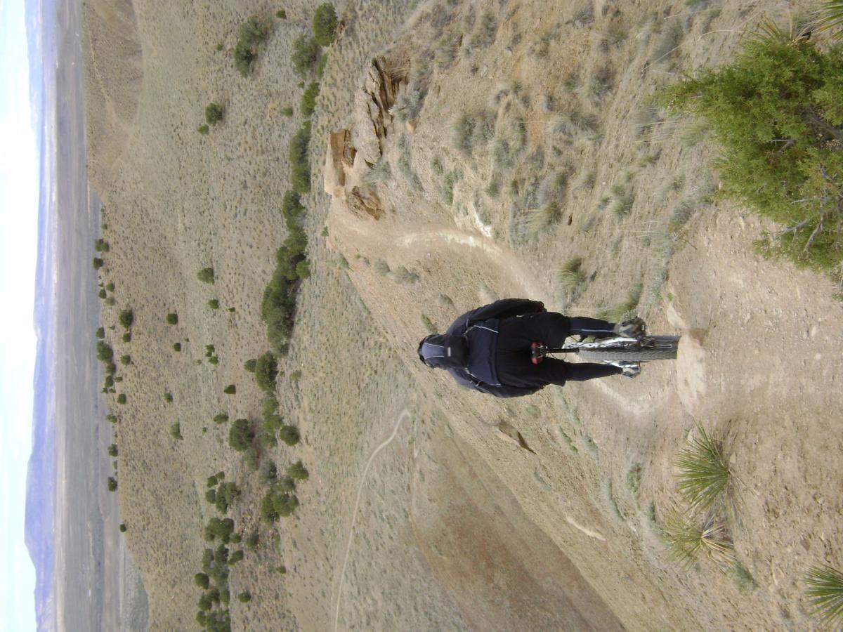 A mountain biker riding along a narrow dirt trail on a hillside, surrounded by sparse vegetation and open terrain. The scene captures the cyclist from behind, highlighting the winding path and expansive landscape in the background, with a cloudy sky above. Joe's Ridge mountain bike trail.