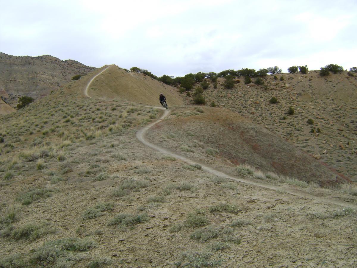 A mountain biker riding along a winding dirt trail on a hilly landscape, surrounded by sparse vegetation under a cloudy sky. Joe's Ridge mountain bike trail.