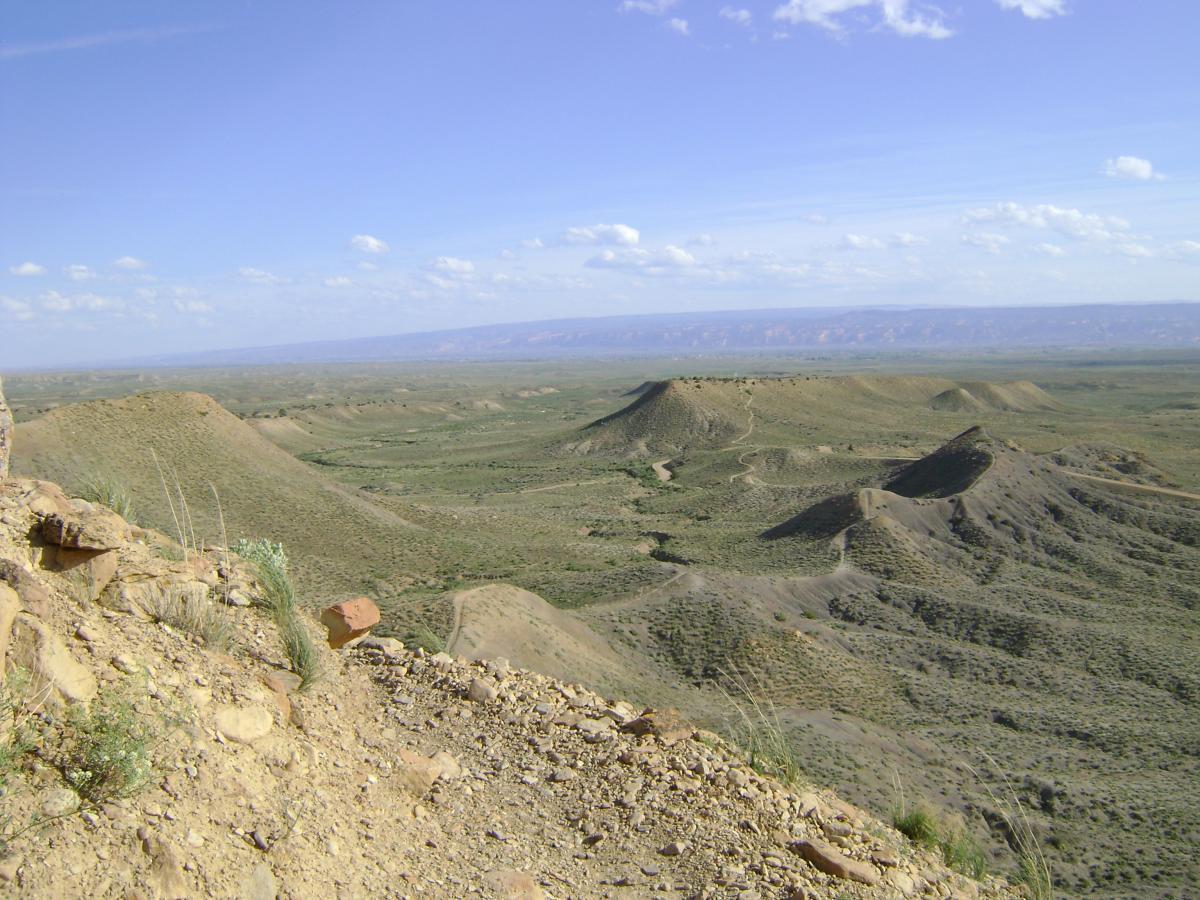 A panoramic view of a vast landscape featuring rolling hills and rugged terrain under a blue sky with scattered clouds. The foreground displays rocky outcroppings and sparse vegetation, while the background reveals additional hills stretching into the distance. The scene conveys a sense of openness and natural beauty. Zippety Do Dah mountain bike trail.