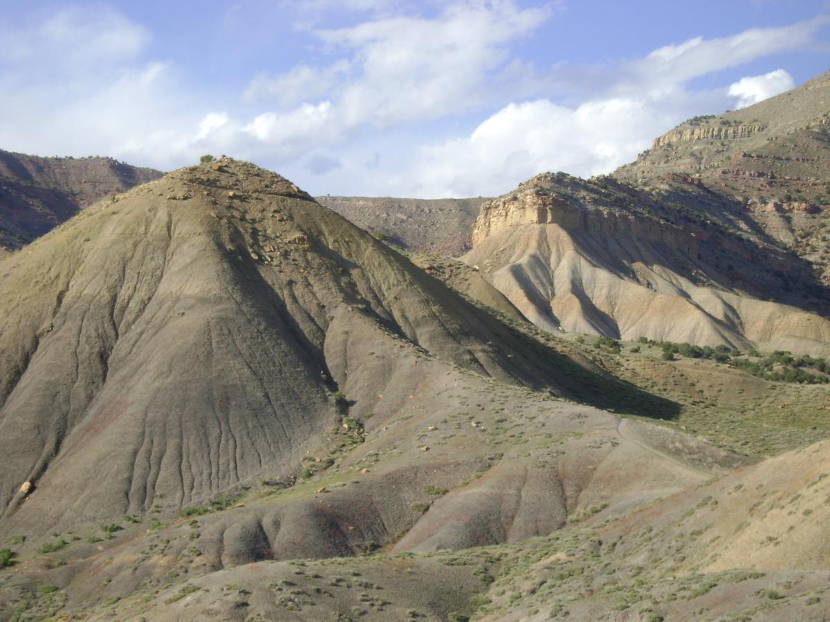 A scenic view of rugged, rolling hills and slopes under a partly cloudy sky. The terrain is characterized by smooth, undulating shapes and subtle color variations, showcasing earthy tones of green, brown, and gray. Small patches of vegetation are visible amidst the rocky landscape. Zippety Do Dah mountain bike trail.