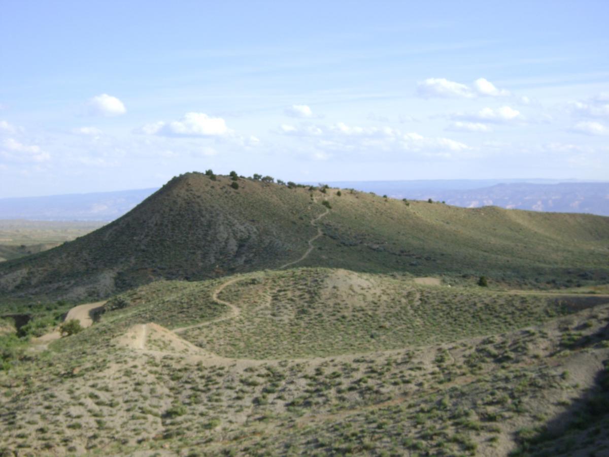 A panoramic view of a gently sloping hill in a sparse, grassy landscape under a blue sky with scattered clouds. A winding dirt path leads up the hillside, surrounded by low shrubs and vegetation. The distant hills and valleys create a sense of depth in the landscape. Zippety Do Dah mountain bike trail.