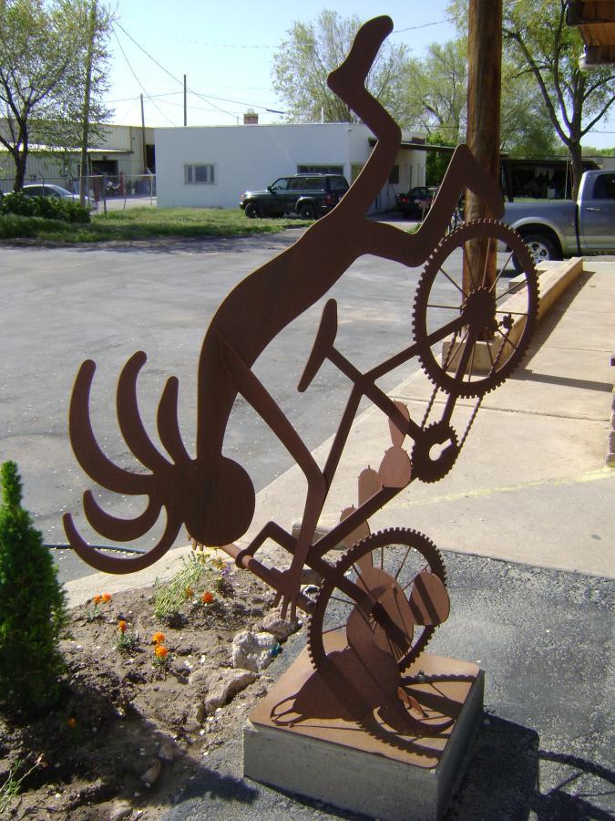 A metal sculpture of a person riding a bicycle on one wheel, showcasing an artistic depiction of balance and action. The figure, crafted from rust-colored metal, has exaggerated features and wild hair, and is positioned in a dynamic pose against a backdrop of grass and a parking area.