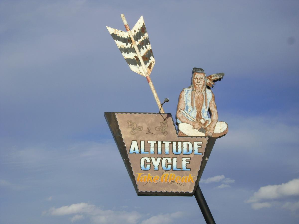 Sign for "Altitude Cycle" featuring a sitting figure in traditional attire and an arrow above it; the background shows a blue sky with clouds.