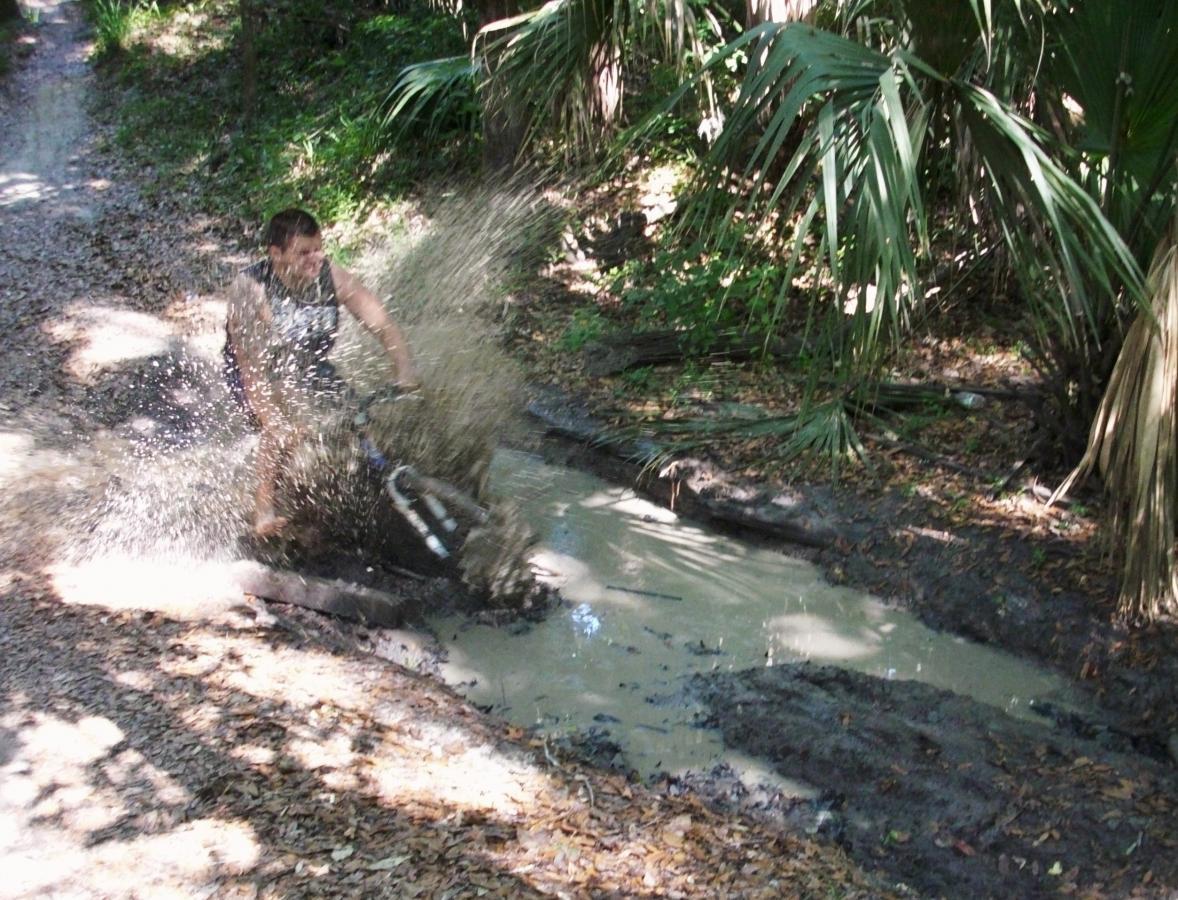 A person riding a bicycle splashes through a muddy puddle on a forest trail, surrounded by green foliage and palm-like plants. The water sprays up around them, creating a dynamic and adventurous scene. Chuck Lennon Park mountain bike trail.