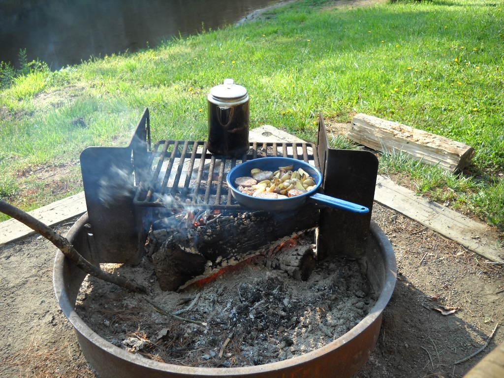 A blue frying pan cooking sliced vegetables over an outdoor campfire, with smoke rising in the background and a coffee pot nearby, set against a grassy landscape with a calm water surface in the distance. New River Trail State Park mountain bike trail.
