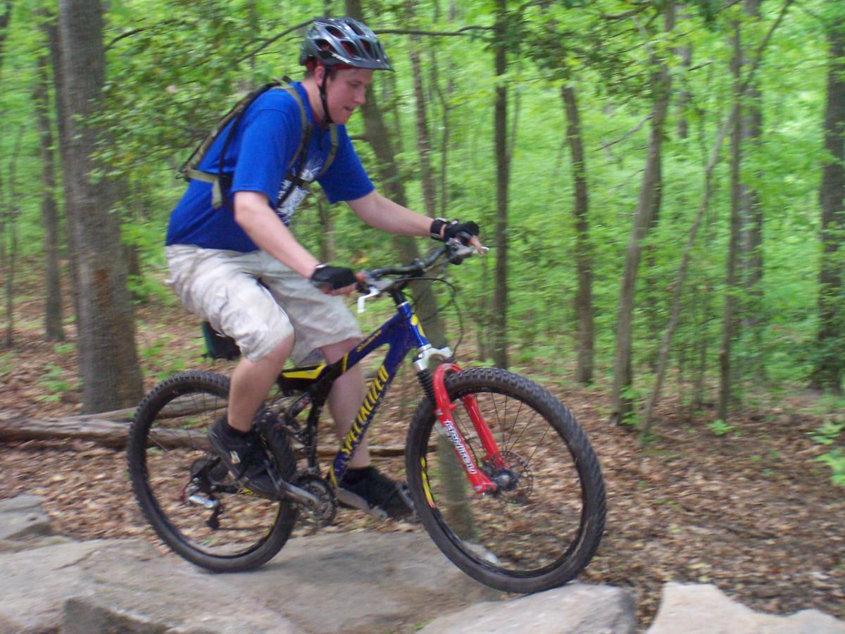 A young man riding a mountain bike over rocky terrain in a wooded area. He is wearing a blue shirt, beige shorts, and a helmet, focusing intently as he navigates the trail surrounded by lush greenery. Powhite Park mountain bike trail.