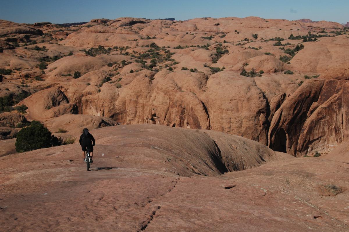 A person riding a mountain bike on rocky terrain, surrounded by a landscape of reddish formations and sparse vegetation under a clear blue sky. The scene captures a sense of adventure and the rugged beauty of the outdoors. Slickrock mountain bike trail.