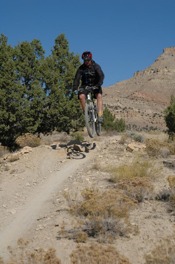 A mountain biker performing a jump on a dirt trail, surrounded by shrubs and rocky terrain, under a clear blue sky. Prime Cut mountain bike trail.