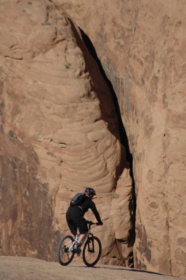 A cyclist riding a mountain bike along a rocky path, with steep, textured canyon walls in the background. The terrain is arid and rugged, showcasing natural rock formations. Slickrock mountain bike trail.