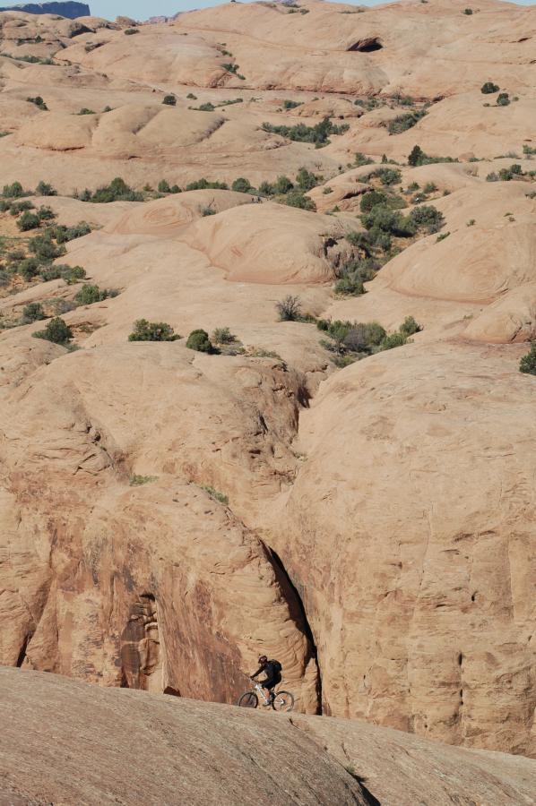 A mountain biker rides along a rocky terrain with beige and reddish rock formations and sparse vegetation in the background. The landscape features smooth, rolling hills, showcasing the natural beauty of the desert environment. Slickrock mountain bike trail.