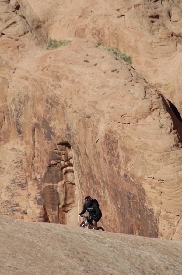 A mountain biker riding on a rocky terrain with a large, textured sandstone formation in the background. The landscape features warm, earthy tones and a clear sky, highlighting the adventurous setting. Slickrock mountain bike trail.