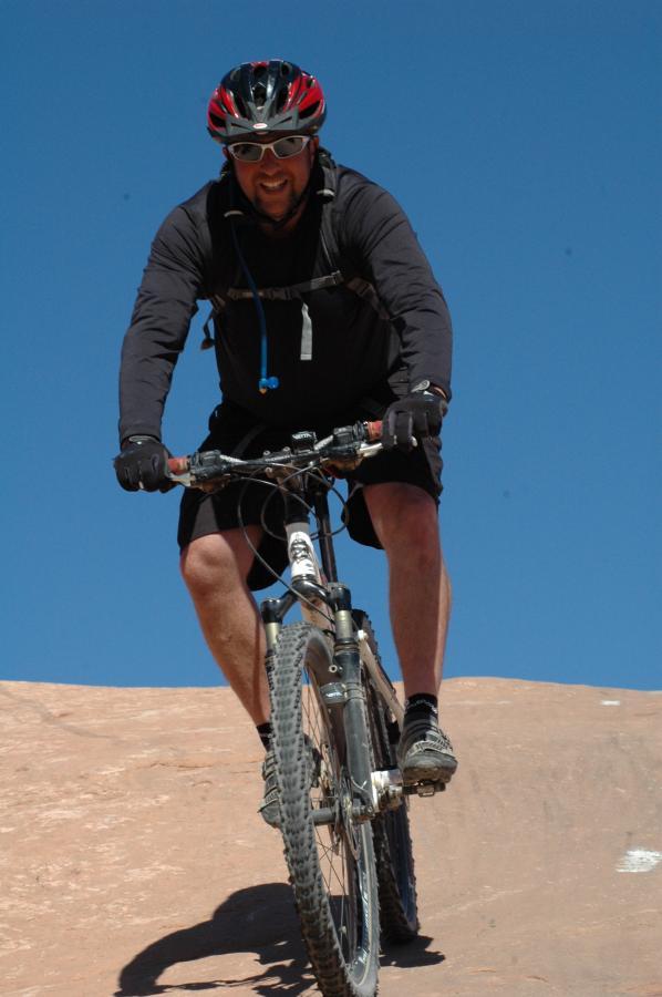 A mountain biker navigating rocky terrain, wearing a black long-sleeve shirt, black shorts, and a red and black helmet. The background features a clear blue sky, emphasizing the outdoor adventure setting. The biker appears focused and determined as they ride downhill. Slickrock mountain bike trail.