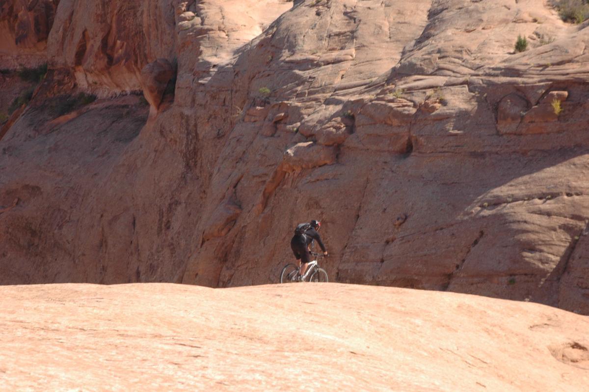A lone mountain biker riding on a rocky, sandy terrain with steep cliffs in the background. The landscape features warm, reddish hues and rugged rock formations under a clear blue sky. Slickrock mountain bike trail.