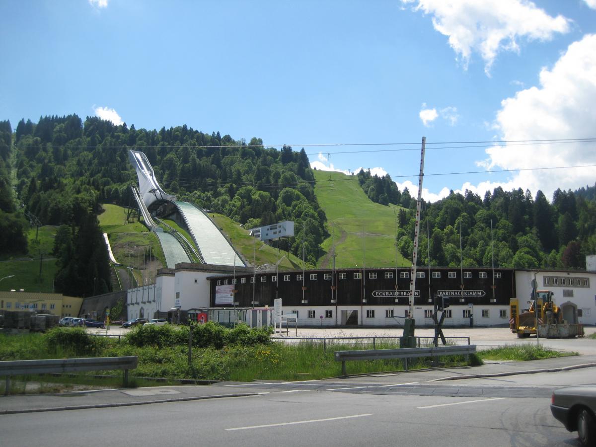 A ski jump ramp rises prominently on a hillside, surrounded by lush green trees under a clear blue sky. In the foreground, a building with a sloped roof and large windows is partially visible, along with a parking area and construction equipment. The landscape conveys a sense of outdoor sport and recreational activity. Garthof Partnachklamm Trail mountain bike trail.