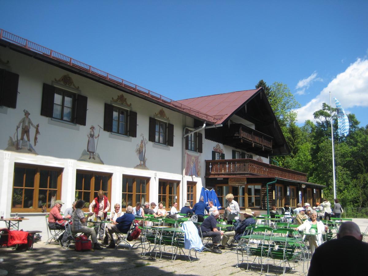 A scenic outdoor dining area at a traditional Alpine lodge, featuring a two-story building with decorative murals on the walls. Guests are seated at green metal tables and chairs, enjoying meals and drinks. The setting is bright and sunny, with a clear blue sky, and surrounded by lush greenery. Garthof Partnachklamm Trail mountain bike trail.