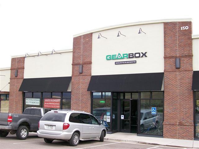 Alt text: Exterior view of a sports equipment store named "Gearbox Mountain Sports," featuring a glass entrance and large awnings. Several parked vehicles are visible in front of the store, with an "Open" sign displayed.