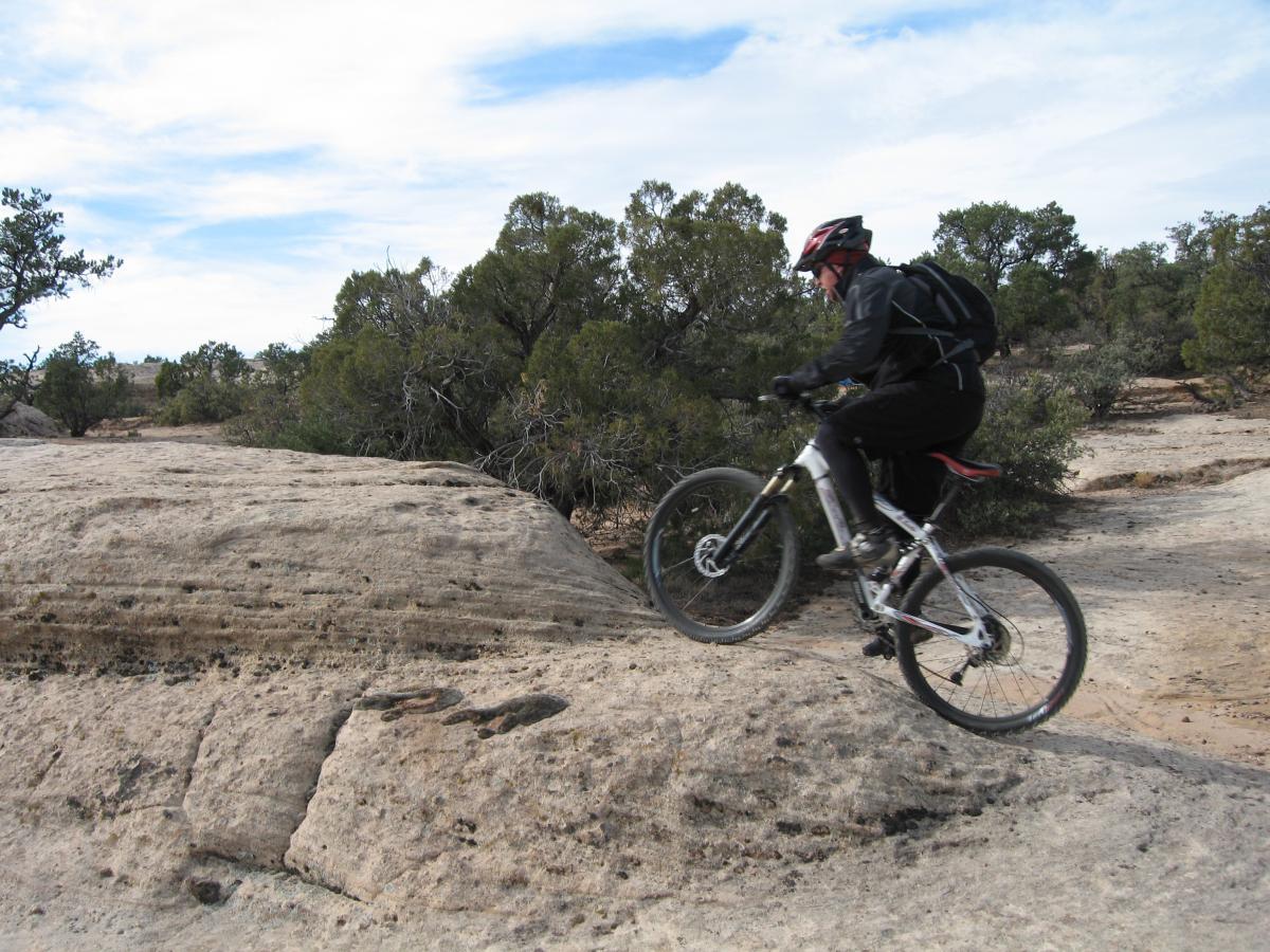 A mountain biker in black gear and a helmet jumps over a rocky terrain, surrounded by sparse vegetation and trees under a blue sky with some clouds. Gooseberry Mesa mountain bike trail.