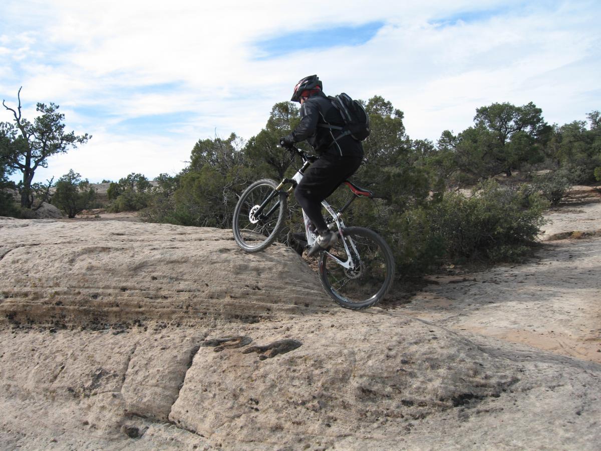 A mountain biker ascends a rocky terrain, showcasing the skill and agility required for off-road cycling. The scene includes a rugged landscape with scattered trees and a blue sky in the background. Gooseberry Mesa mountain bike trail.