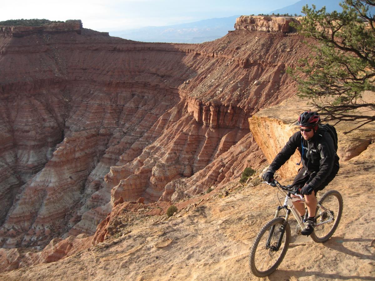 A mountain biker rides along a rocky, cliffside trail with layered red rock formations and a canyon in the background. The cyclist wears a helmet and gear, navigating the challenging terrain during a clear day. Gooseberry Mesa mountain bike trail.