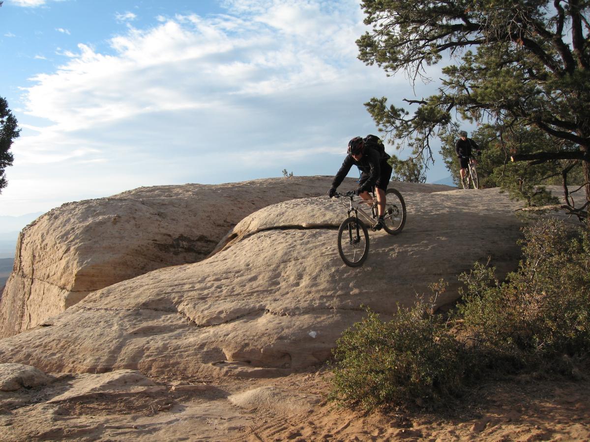 A mountain biker navigating a rocky terrain with a smooth, large rock formation under a clear blue sky, while another biker trails behind on a nearby path. The surrounding area features sparse vegetation and trees. Gooseberry Mesa mountain bike trail.