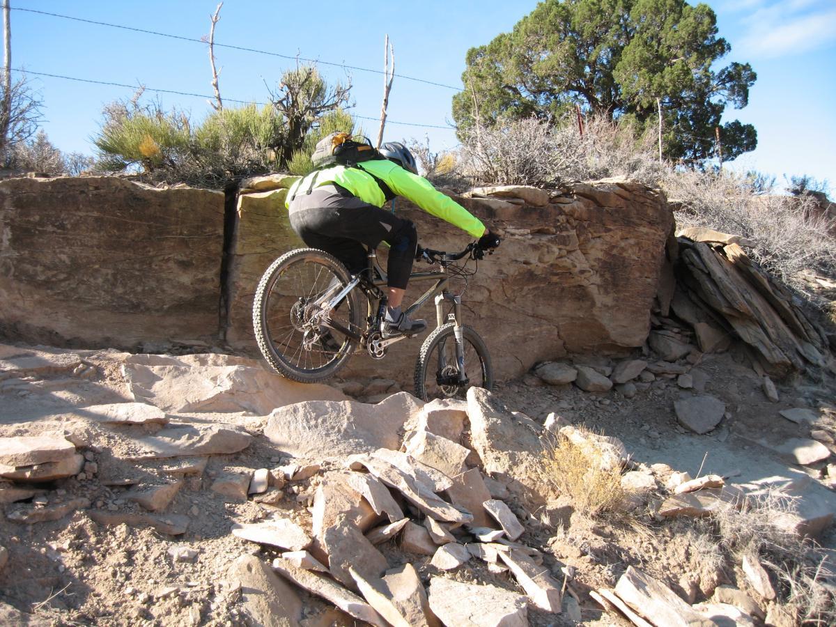 A mountain biker in a bright yellow and black jacket navigates rocky terrain on a mountain bike, with a steep rock face in the background and sparse vegetation around. J.E.M. Trail mountain bike trail.