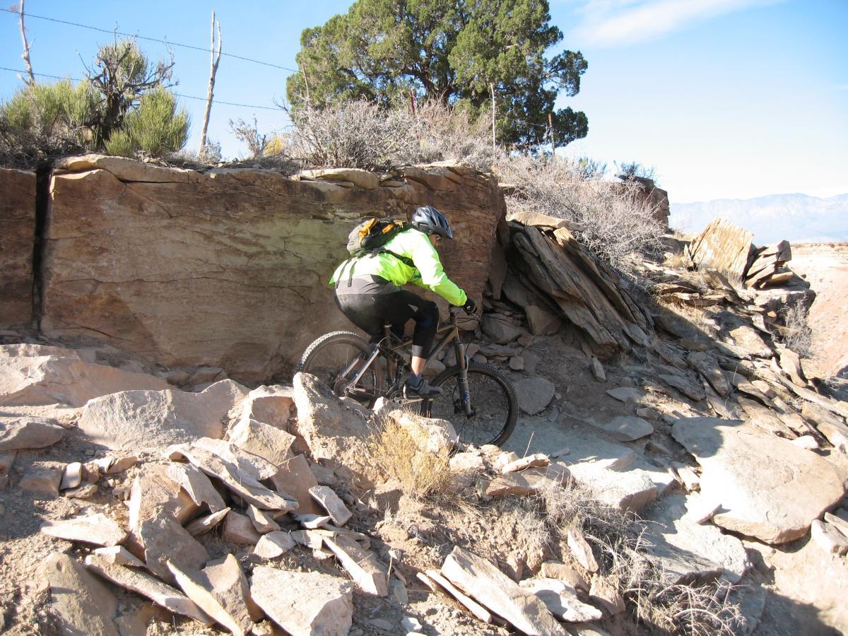 A mountain biker in a bright green jacket navigates a rocky trail surrounded by sparse vegetation and large rock formations, under a clear blue sky. J.E.M. Trail mountain bike trail.