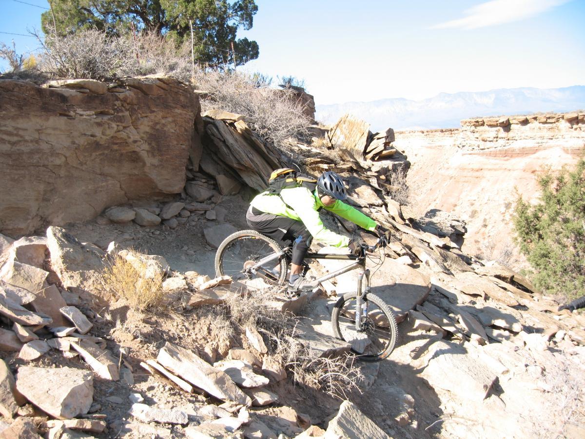 A mountain biker navigating a rocky terrain on a narrow trail, with steep cliffs and distant mountains visible in the background. The rider is wearing a helmet and a bright green jacket, focused on the challenging path ahead. Sagebrush and other vegetation are present alongside the trail. J.E.M. Trail mountain bike trail.