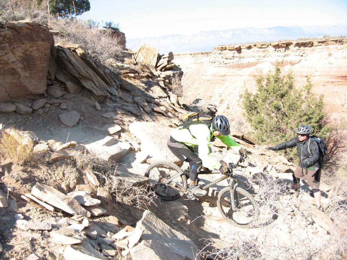 A mountain biker navigating a rocky trail with steep terrain, while another person stands nearby, offering assistance. The background features a rugged landscape and distant mountains under a clear sky. J.E.M. Trail mountain bike trail.