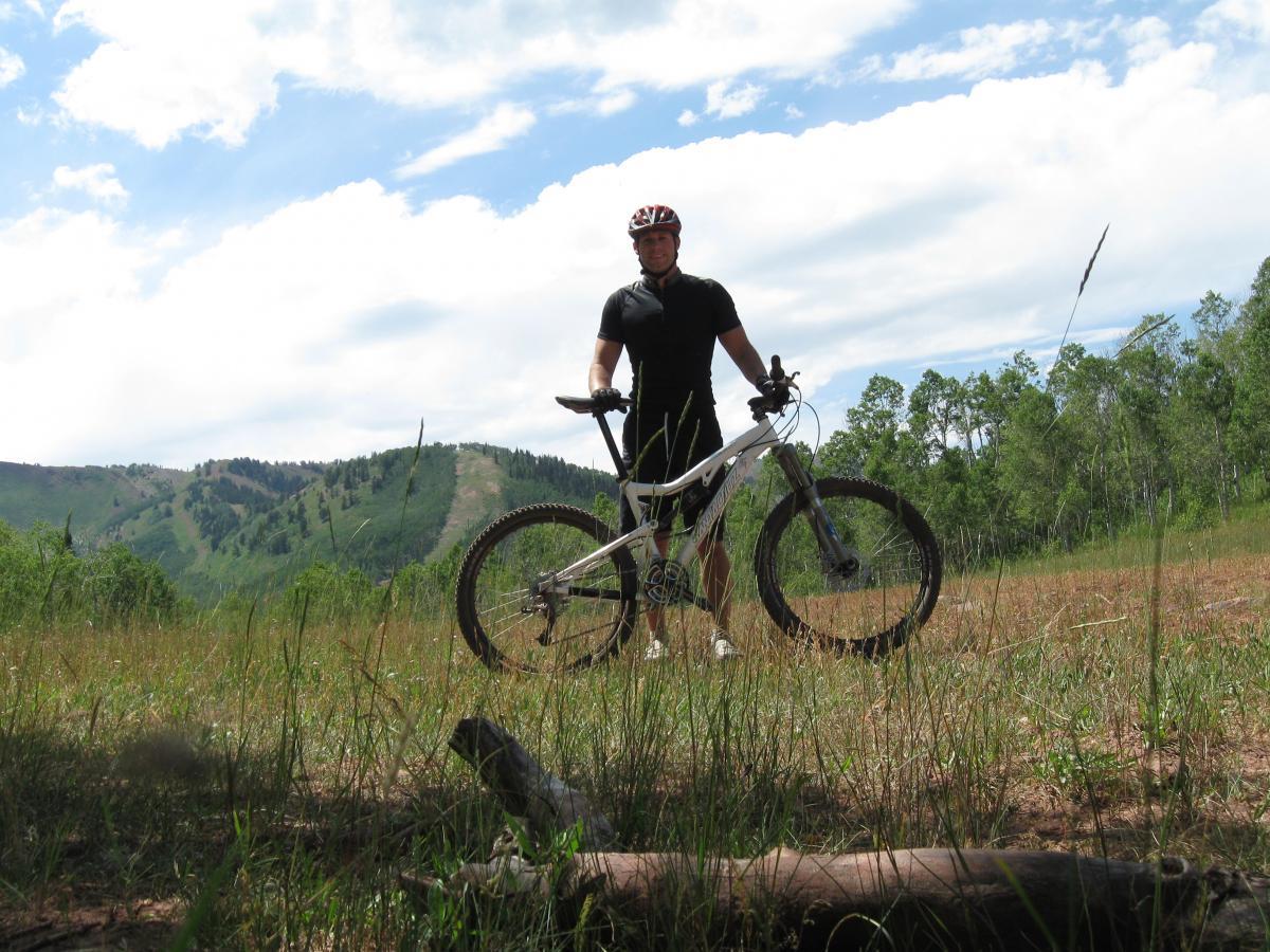 A person stands with a mountain bike in an open field surrounded by green hills and trees, under a partly cloudy sky. The individual is wearing a helmet and athletic clothing, ready for cycling in a natural outdoor setting. Mid Mountain mountain bike trail.