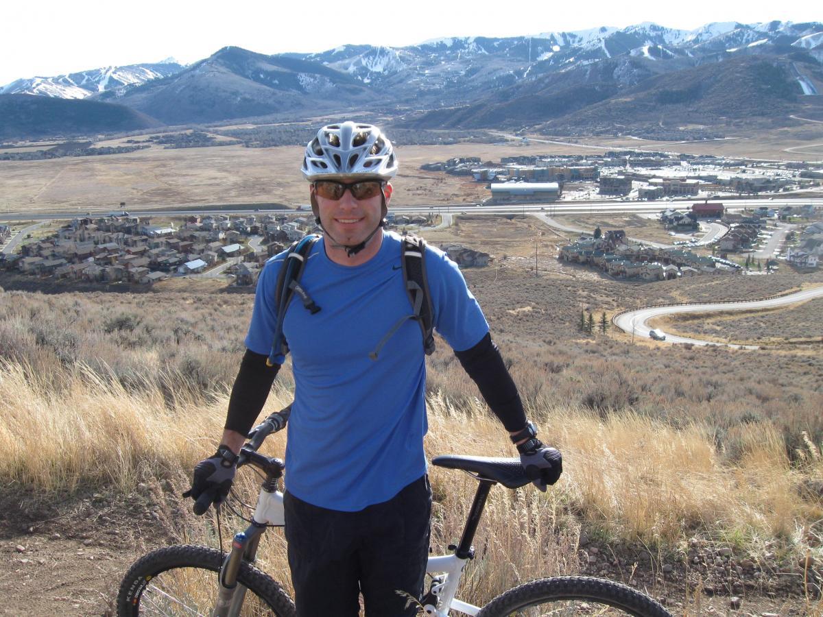 A mountain biker posing with his bike on a hillside, overlooking a valley with a small town and mountains in the background. The biker is wearing a blue shirt, gloves, sunglasses, and a helmet, with patches of grass and shrubs around him. Snow-capped mountains are visible in the distance under a clear sky. Glenwild mountain bike trail.