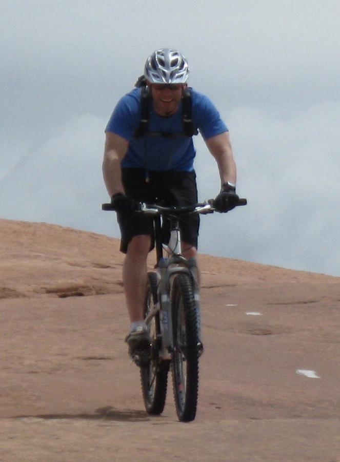 A cyclist in a blue shirt and black shorts rides a mountain bike on a rocky trail, wearing a helmet and gloves, with a cloudy sky in the background. Slickrock mountain bike trail.