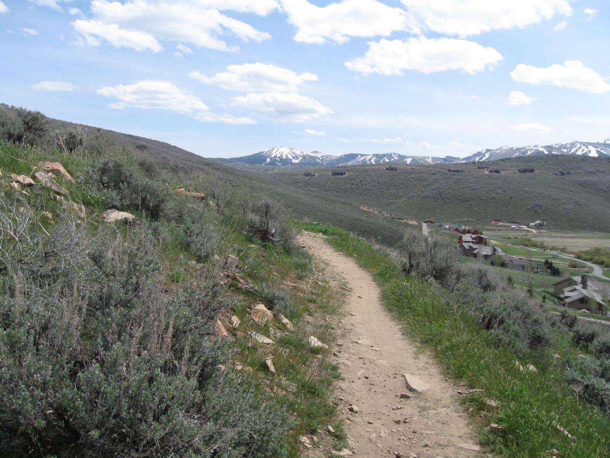 A dirt trail winding through greenery and shrubs, leading through a hilly landscape with snow-capped mountains in the distance under a partly cloudy sky. In the foreground, the trail is surrounded by rocks and grasses, while a small settlement is visible further down the valley. Round Valley mountain bike trail.