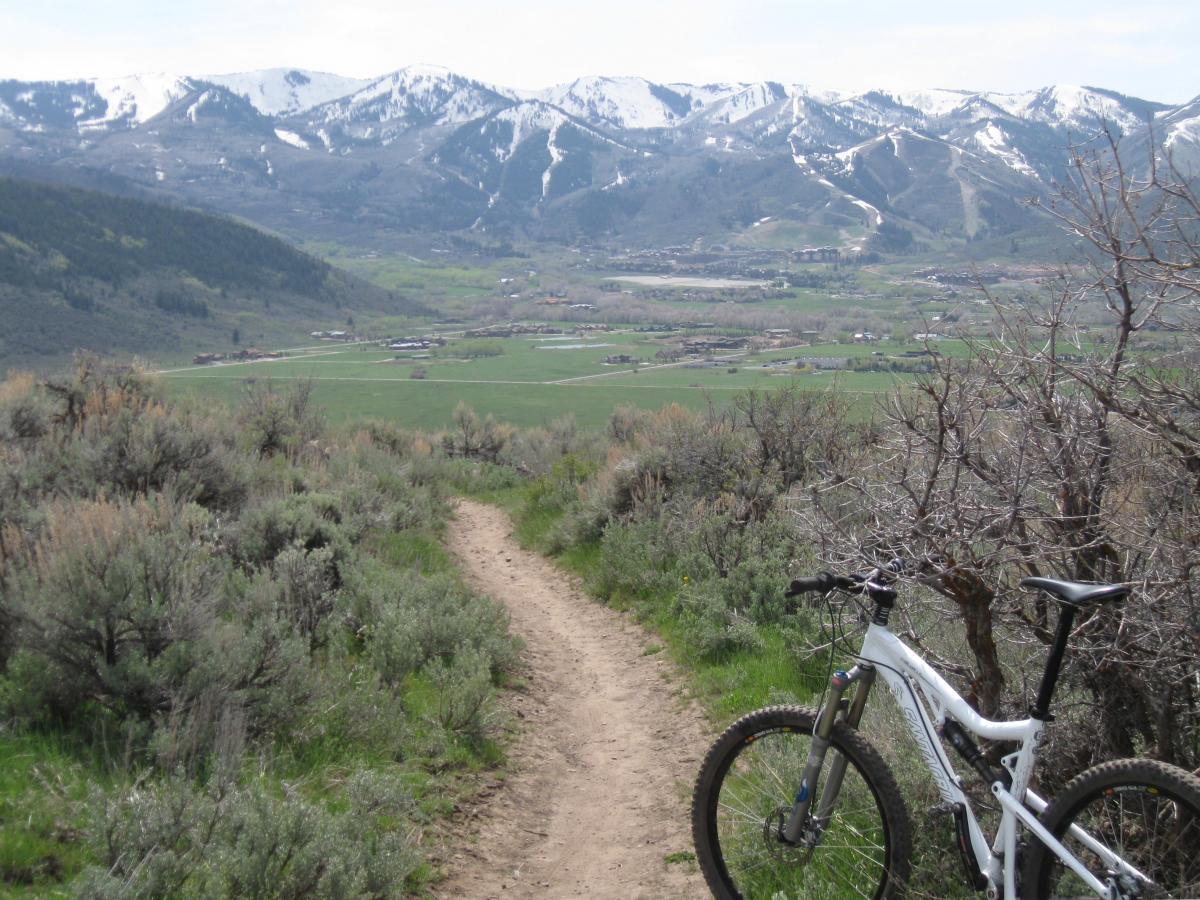 A mountain biking trail winding through green shrubbery, with a white mountain bike leaning on a bush in the foreground. In the background, snow-capped mountains rise above a lush valley dotted with patches of grass and small buildings under a clear blue sky. Round Valley mountain bike trail.