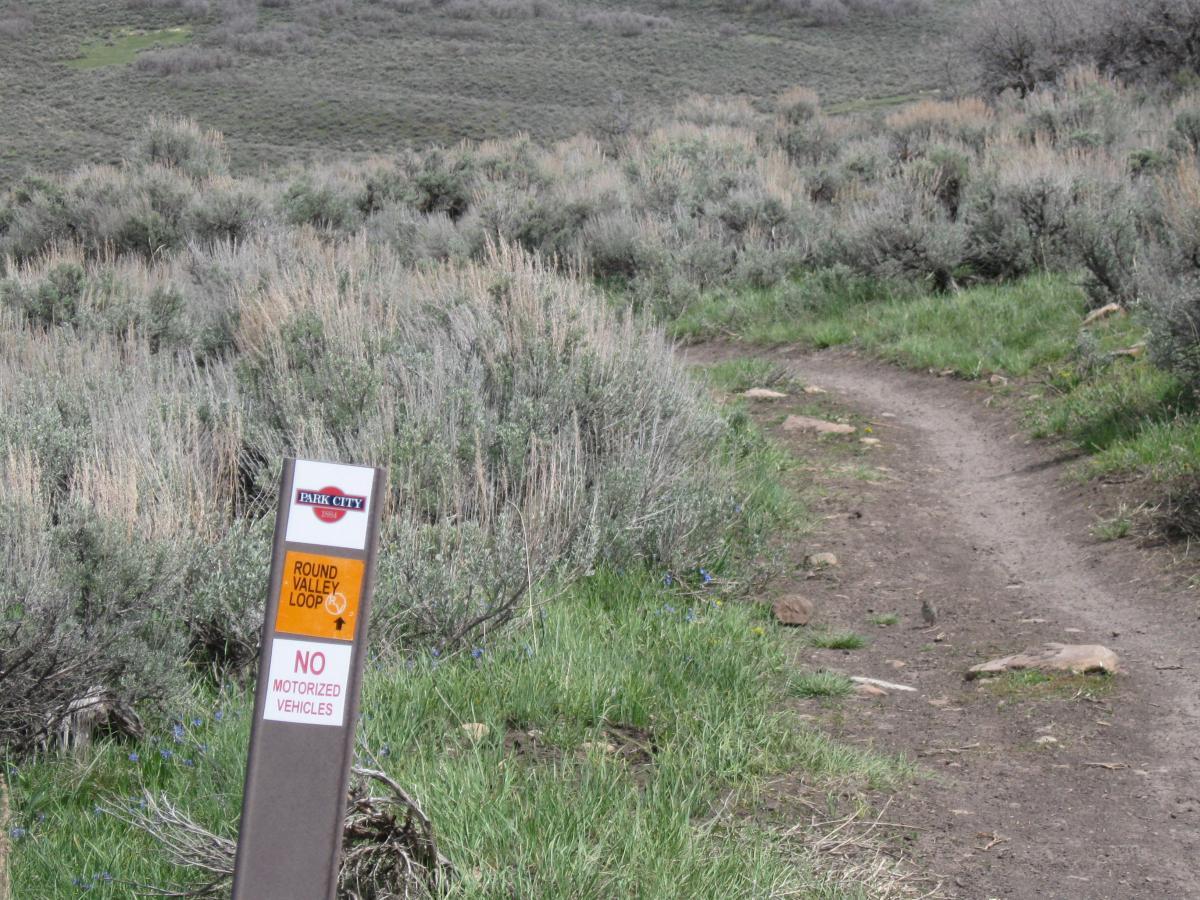 Sign at the Round Valley Loop trailhead in Park City, indicating no motorized vehicles allowed, surrounded by green grass and shrubs. A dirt path winds away from the sign into the natural landscape. Round Valley mountain bike trail.
