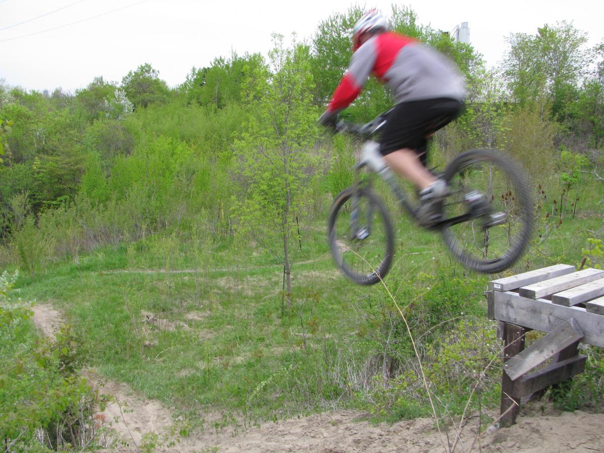 A mountain biker in a gray and red outfit jumps off a wooden ramp, soaring through the air above a grassy, green landscape with trees and shrubs in the background. Don Valley mountain bike trail.