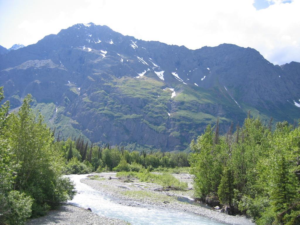 A scenic view of a winding river flowing through lush greenery, with towering mountains in the background, partially covered in snow under a bright blue sky. Eklutna Lake Lakeside Trail mountain bike trail.