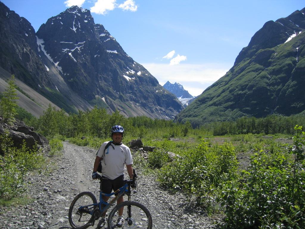 A person wearing a helmet and cycling gear stands beside a mountain bike on a rocky trail in a mountainous landscape. The scene features steep, snow-capped mountains in the background under a clear blue sky, with lush greenery and small bushes lining the path. Eklutna Lake Lakeside Trail mountain bike trail.