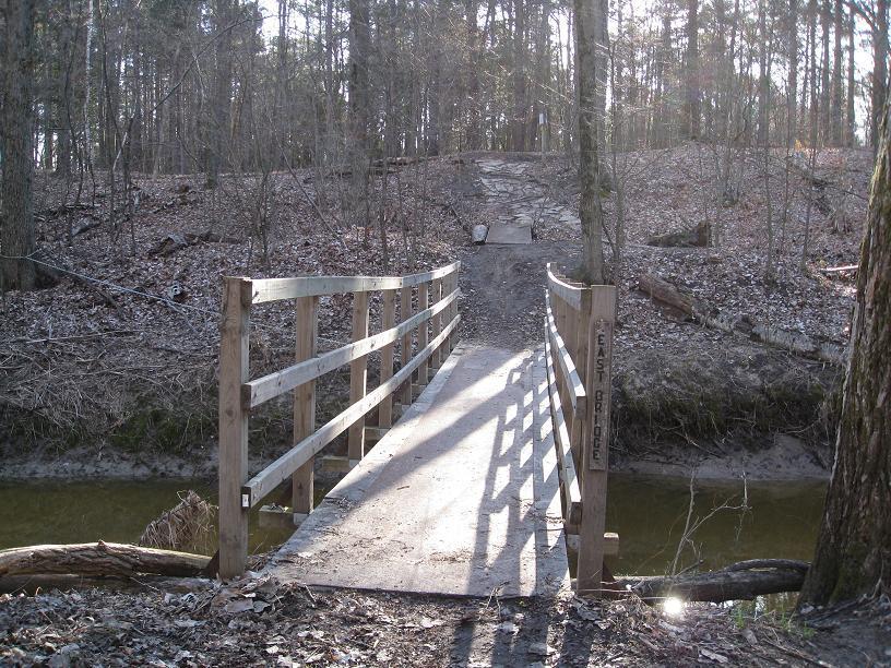 A wooden footbridge spans a small creek, leading into a wooded area with bare trees and a gravel path. The sunlight casts shadows on the bridge, and leaf litter covers the ground in the forest. Midland City Forest mountain bike trail.