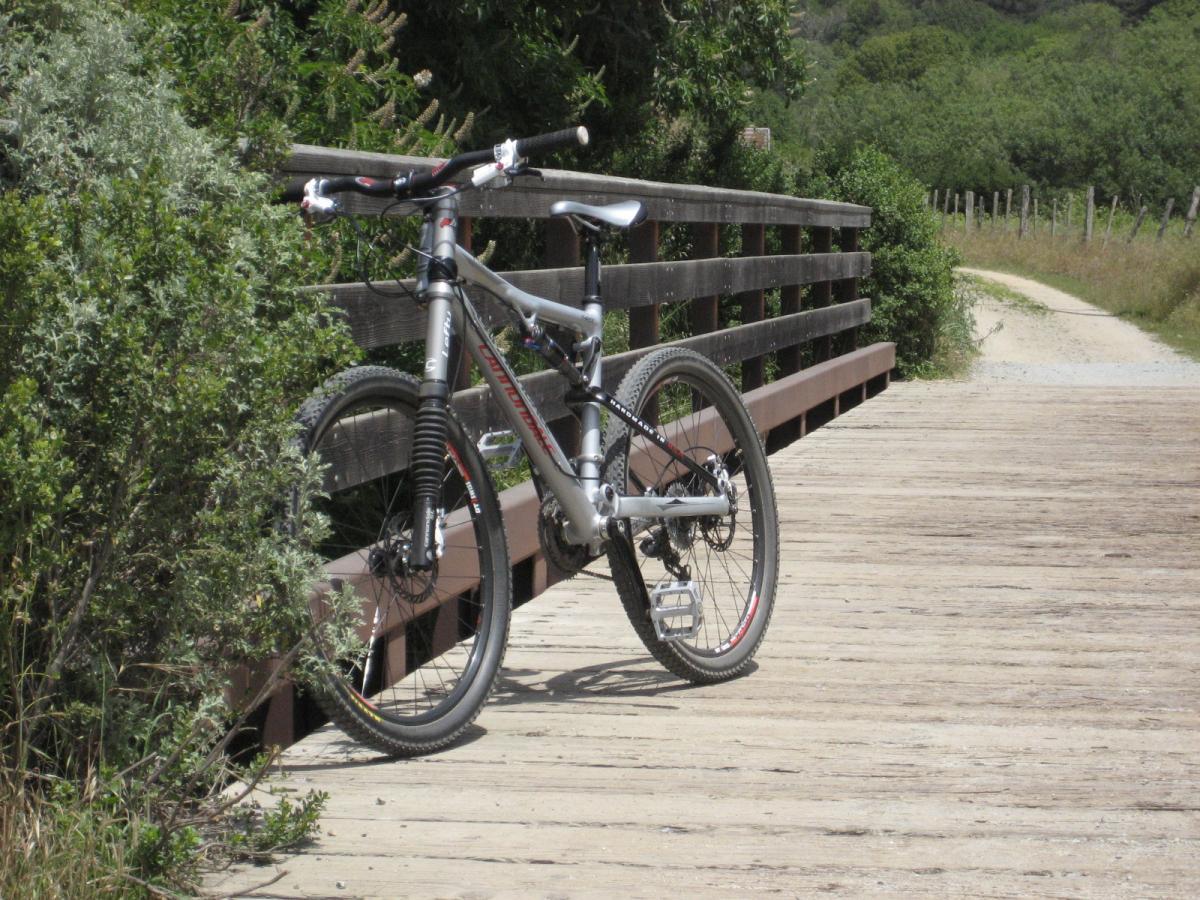 A mountain bike resting on a wooden bridge surrounded by greenery, with a dirt path visible in the background. Wilder Ranch State Park mountain bike trail.