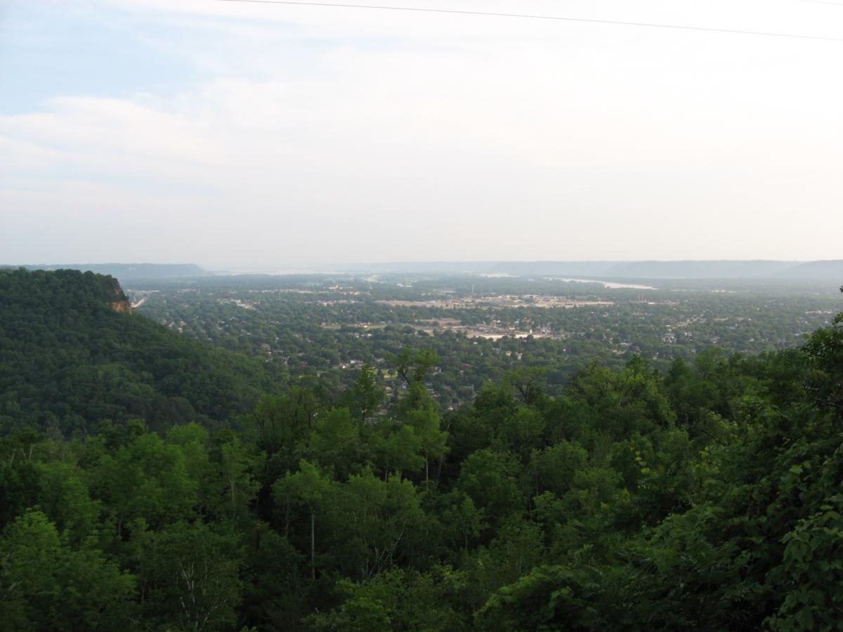 A panoramic view of a lush green landscape with rolling hills and a river in the distance, under a cloudy sky. The foreground features dense trees, while the middle ground showcases a town nestled in the valley, extending towards the horizon. Bluff Trails mountain bike trail.
