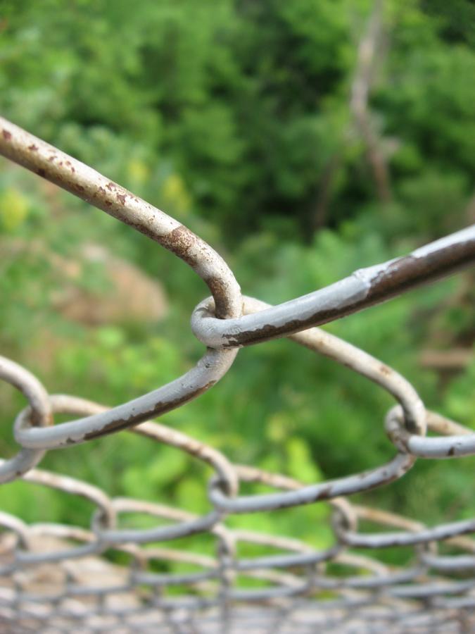 Close-up view of a rusty chain-link fence, with focus on the intertwined metal links against a blurred green background of foliage. Bluff Trails mountain bike trail.