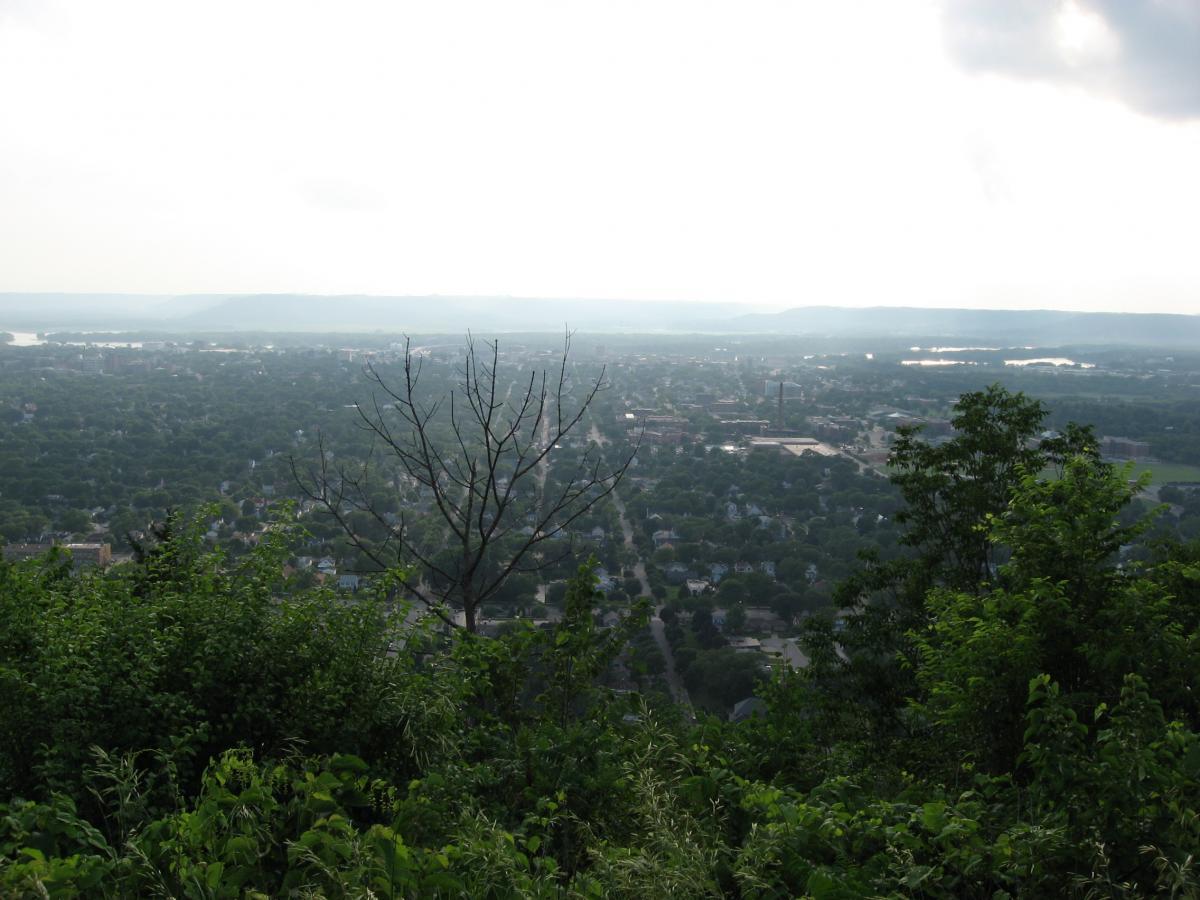 A panoramic view of a town nestled in a green valley, with a foreground of lush foliage and a leafless tree, under a hazy sky. The landscape stretches into the distance, revealing hills and a river visible beyond the town's outskirts. Bluff Trails mountain bike trail.